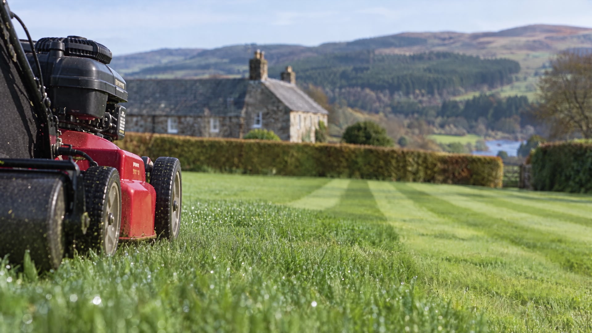 A petrol roller mower laying a straight stripe across a large lawn in front of a Borders stone house